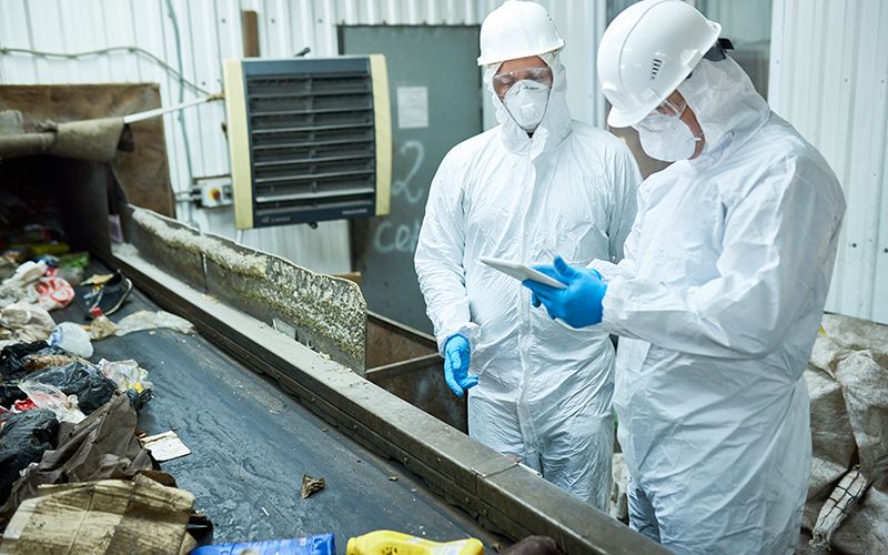 Portrait of two workers  wearing biohazard suits working at waste processing plant sorting trash and using digital tablet standing by conveyor belt, copy space